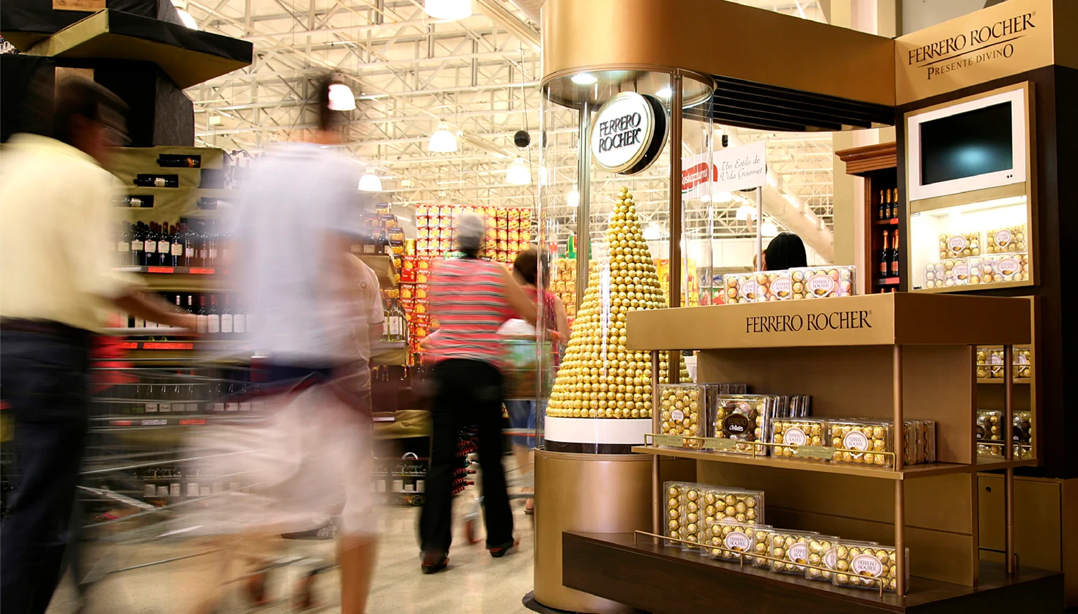 Quiosque Ferrero Rocher dourado em supermercado, com clientes em movimento e torre de chocolates.